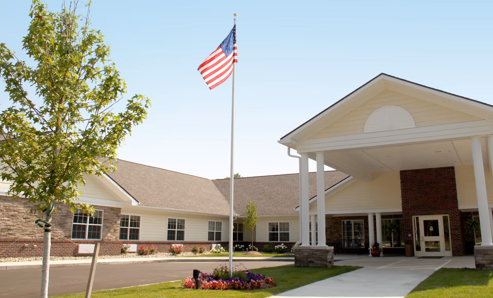 Front entrance of a single-story senior living facility with a covered portico, an American flag on a flagpole, windows, and landscaped grounds.