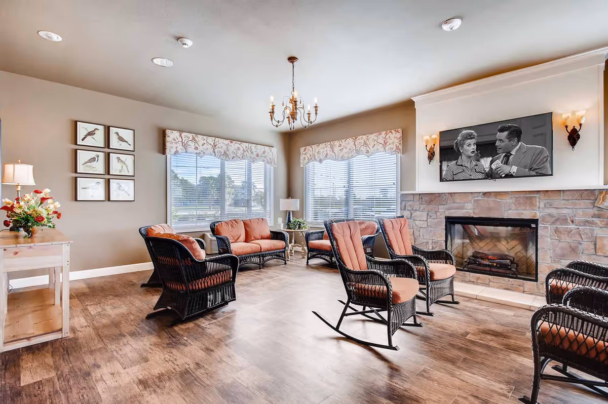 A cozy living room with wicker furniture including rocking chairs and sofas with orange cushions arranged around a stone fireplace. Above the fireplace is a wall-mounted TV displaying a black and white show. The room has large windows with floral valances letting in natural light, a chandelier hanging from the ceiling, framed bird artwork on one wall, and a wooden side table with a lamp and flower arrangement.