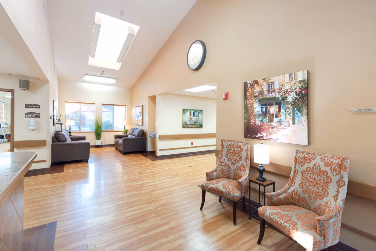A bright and spacious common area in a senior living facility with wooden flooring, two patterned armchairs with a small table and lamp between them, a large wall clock, and a colorful painting on the wall. In the background, there are two dark leather sofas near windows with blinds, potted plants, and soft lighting from lamps.
