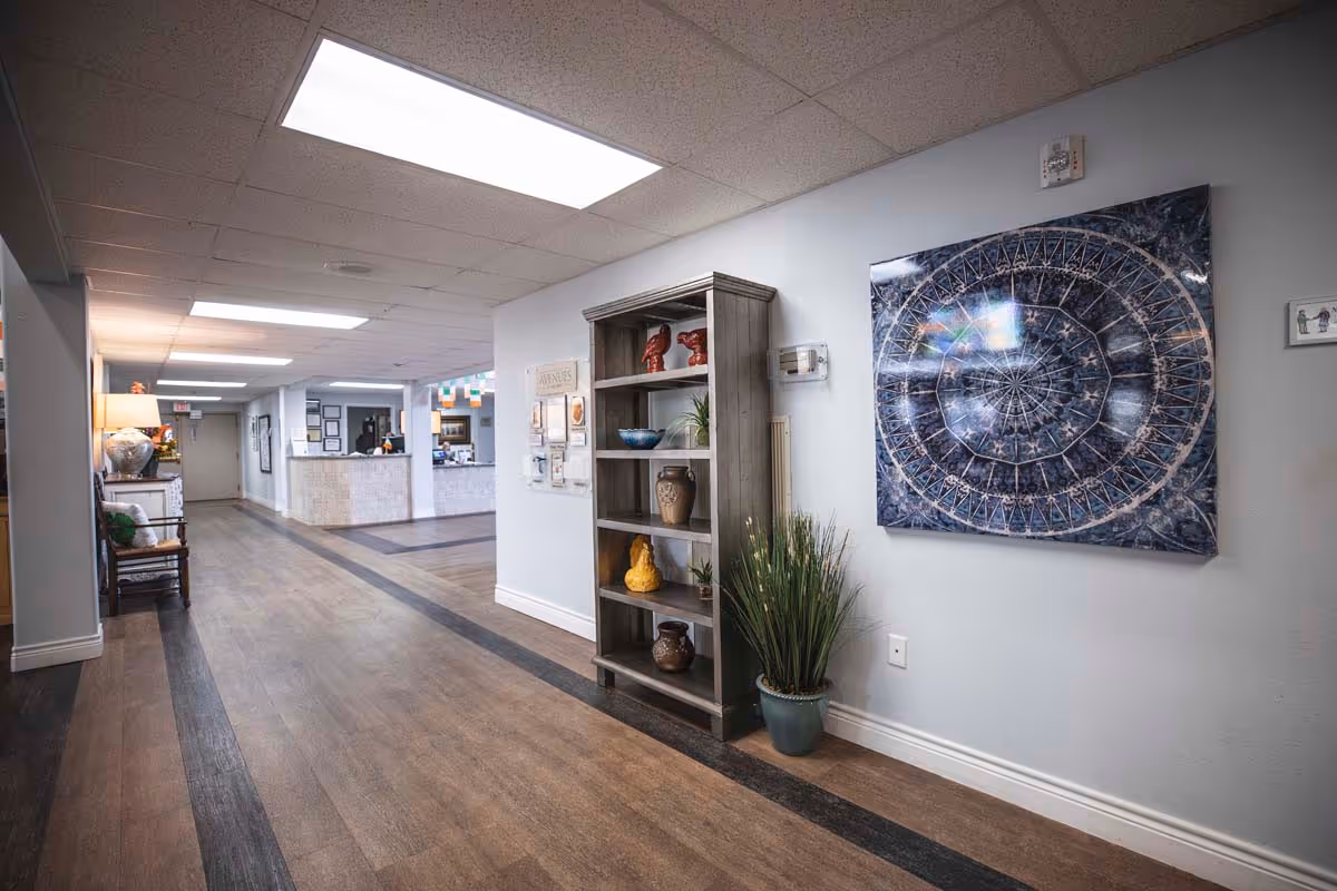 Interior hallway of The Grove on Forest Lane facility with wood flooring and a decorative dark stripe pattern. On the right wall, there is a tall wooden shelf with various decorative items and a potted plant beside it. Above the plant, a large circular abstract artwork is mounted on the wall. The hallway leads to a reception desk area in the background with bright overhead lighting.