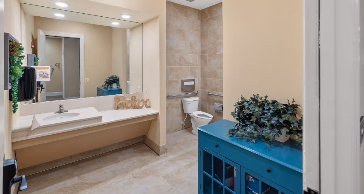 A clean and spacious bathroom with beige tiled floors and walls. There is a large mirror above a white sink countertop with a wooden 'Relax' sign on it. A white toilet with grab bars is visible in the corner. A blue cabinet with a green leafy plant on top is positioned near the entrance. The room is well-lit with a ceiling light.