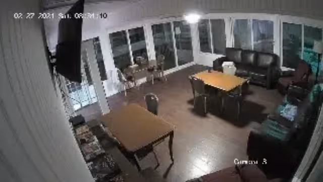 Interior view of a common area with multiple tables and chairs, a black leather couch, and large windows along the walls. The room has wooden flooring and appears to be well-lit with overhead lighting.
