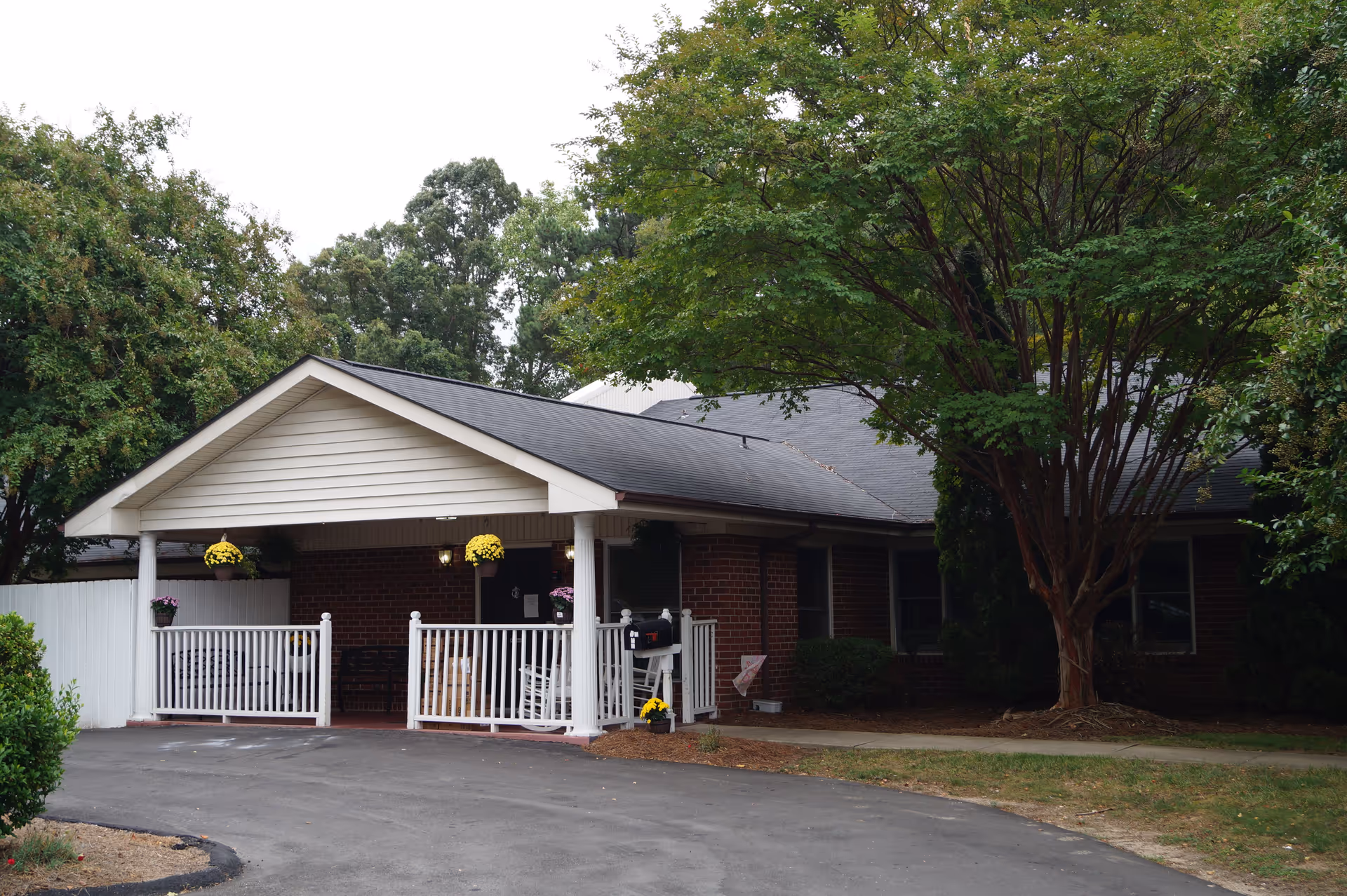 Exterior view of a single-story brick building with a covered porch featuring white railings and columns. The porch has hanging flower pots with yellow flowers and white rocking chairs. There is a large tree to the right of the porch and greenery surrounding the building.