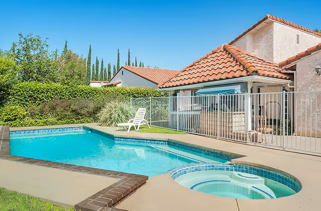 Outdoor pool area at a senior living facility with a hot tub, lounge chairs, and fencing.