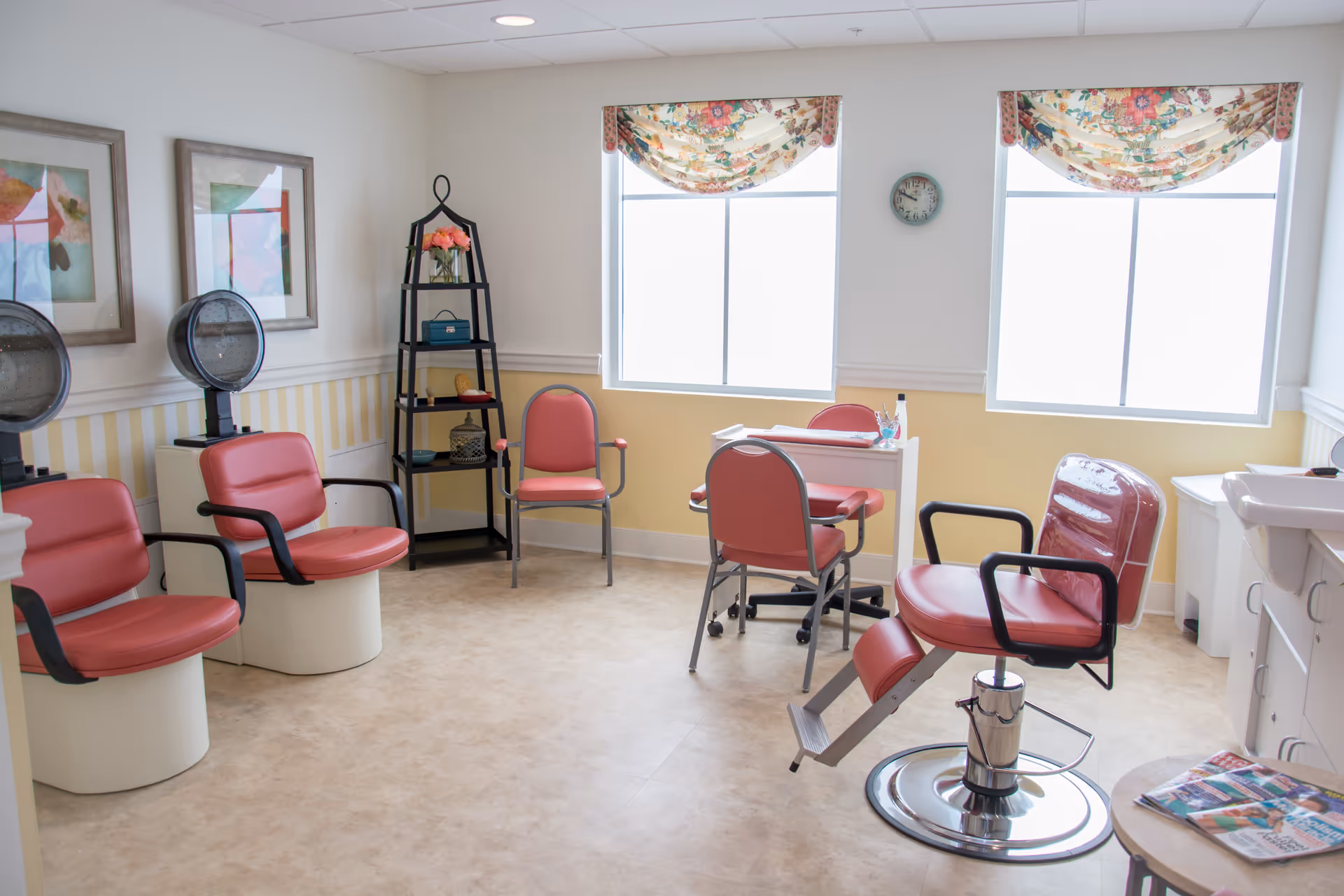 A bright salon room with red salon chairs, hair dryers, a small desk with two chairs, a black shelving unit with decorative items, two large windows with floral valances, and framed artwork on the wall.