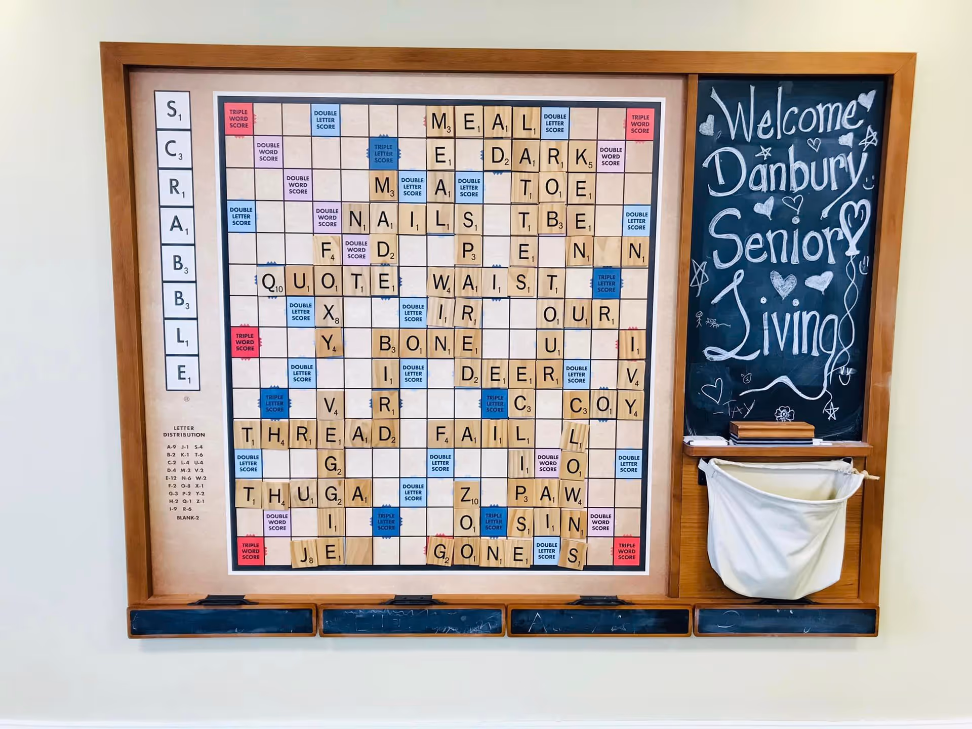 A large wall-mounted Scrabble game board with wooden letter tiles forming various words. To the right of the board is a chalkboard with the handwritten message 'Welcome Danbury Senior Living' decorated with hearts and stars. Below the chalkboard is a small white cloth pouch attached to the wooden frame.