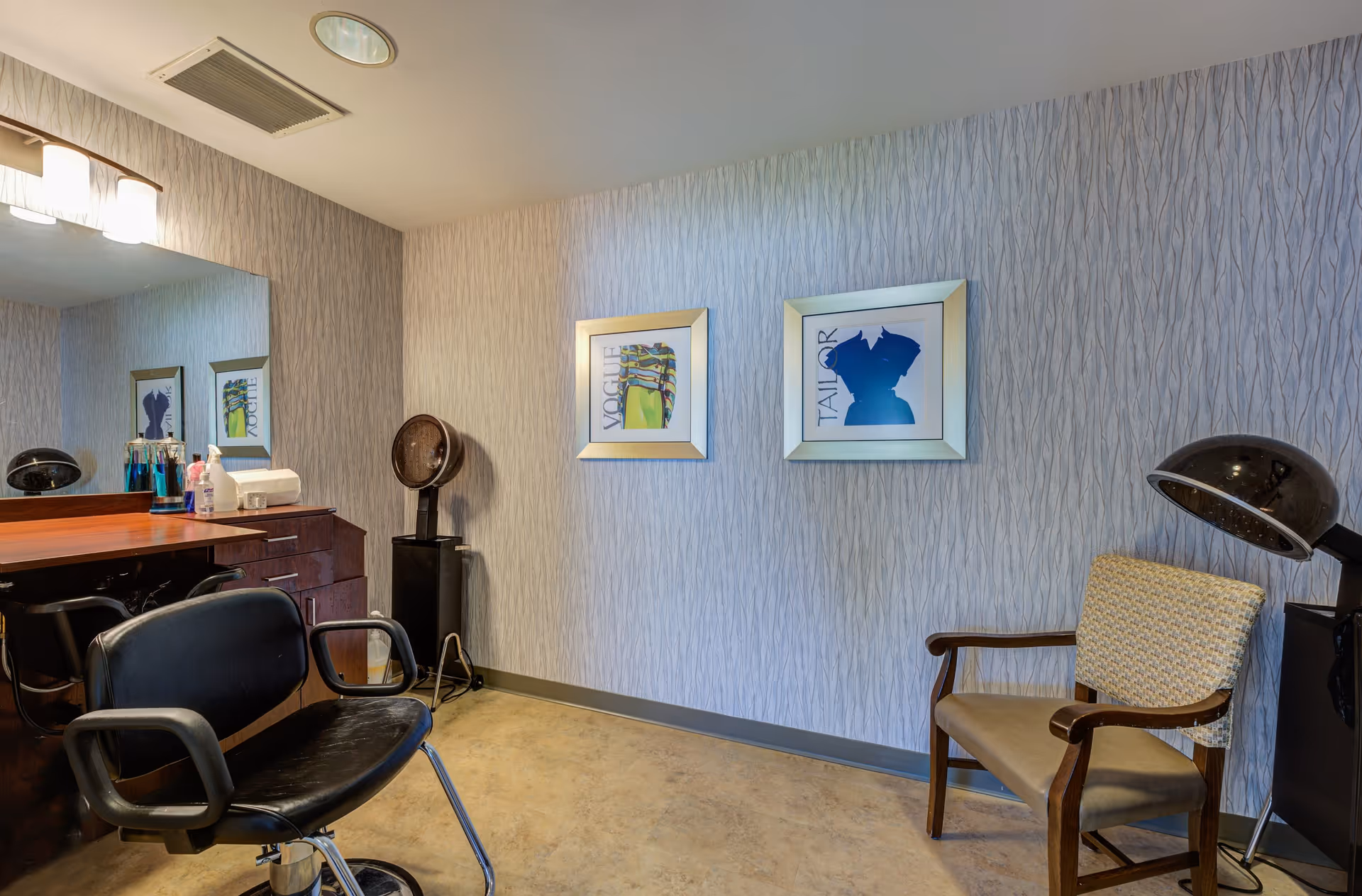 Interior view of a small salon or hair care room with a black salon chair in front of a wooden counter with a large mirror. On the counter are various hair care products and a tissue box. There is a hair dryer hood on a stand near a beige armchair with wooden armrests. The walls have a textured pattern with two framed fashion-themed pictures hanging.