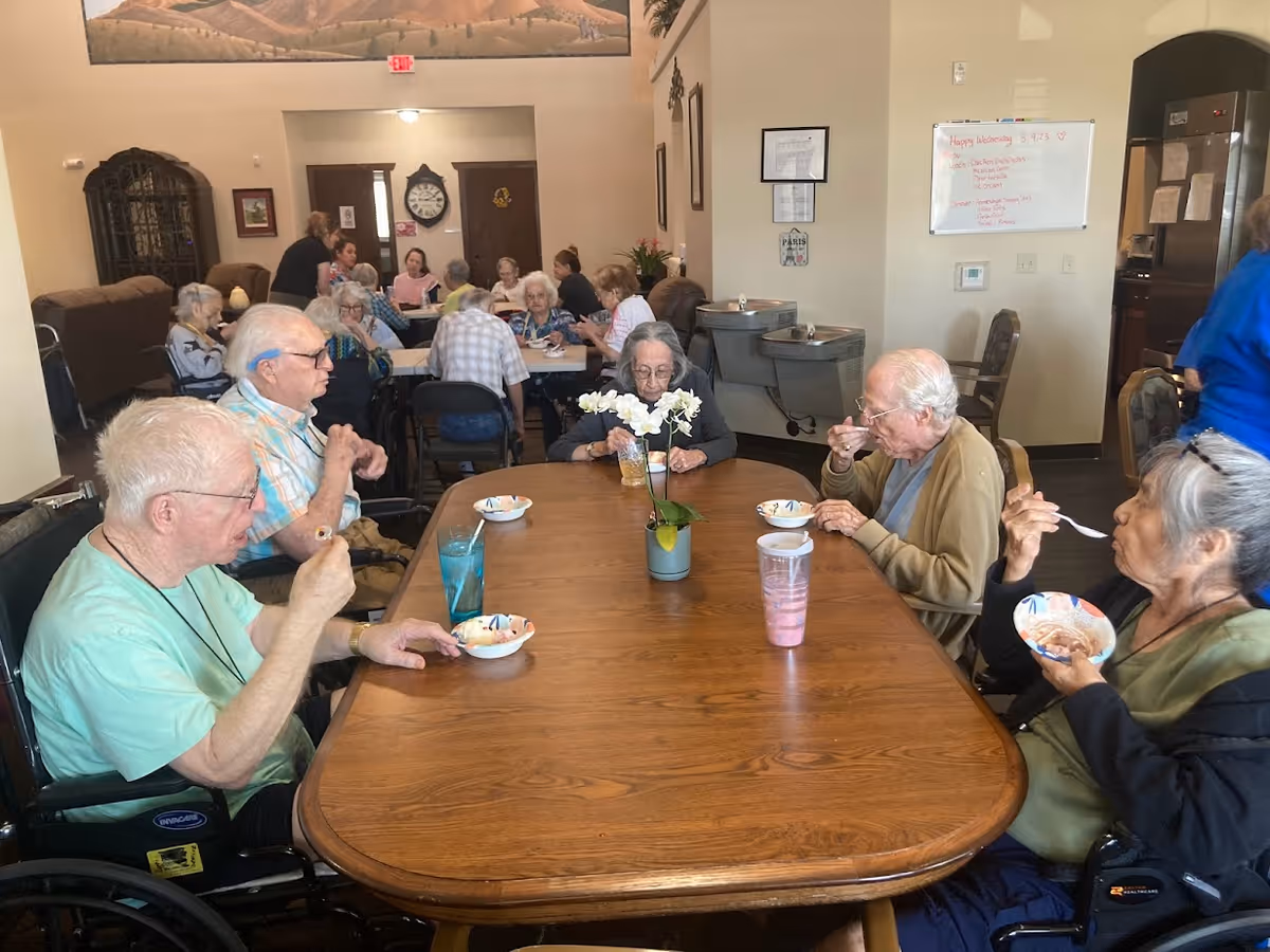 A group of elderly people sitting around a wooden dining table eating from bowls with spoons in a communal dining area. There are more elderly individuals seated at tables in the background, and a whiteboard with writing is visible on the wall. The room has a warm, social atmosphere with comfortable seating and a decorative plant on the table.
