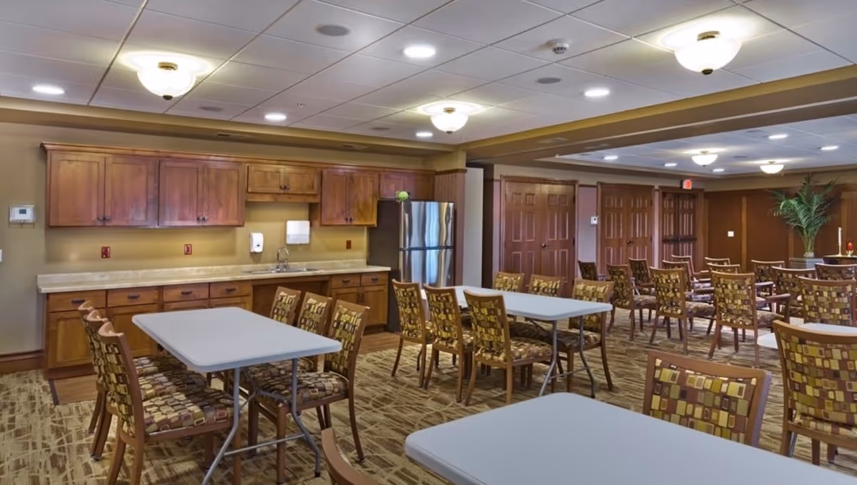 A spacious dining room with multiple tables and chairs arranged neatly. The room features wooden cabinets, a stainless steel refrigerator, and a countertop with a sink. The ceiling has recessed lighting and decorative light fixtures. There is a potted plant in the corner and several closed wooden doors along the back wall.