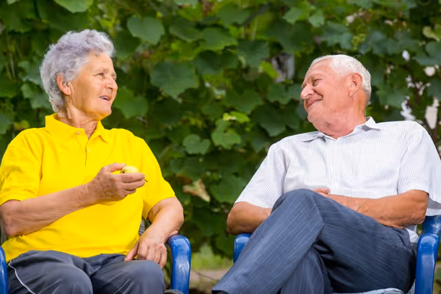 An elderly woman in a yellow shirt and an elderly man in a white striped shirt sitting outdoors on blue chairs, smiling and engaging in conversation with green foliage in the background.