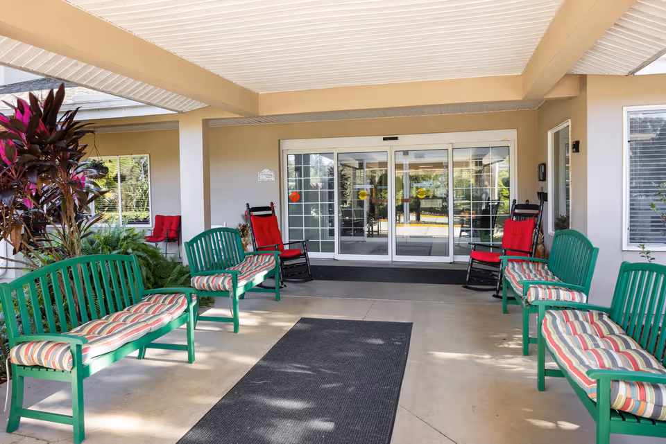 Covered entrance area of a senior living facility with green benches that have striped cushions and red cushioned rocking chairs. The entrance has automatic sliding glass doors and is surrounded by beige walls and windows. There are plants on the left side and a black mat on the floor leading to the doors.