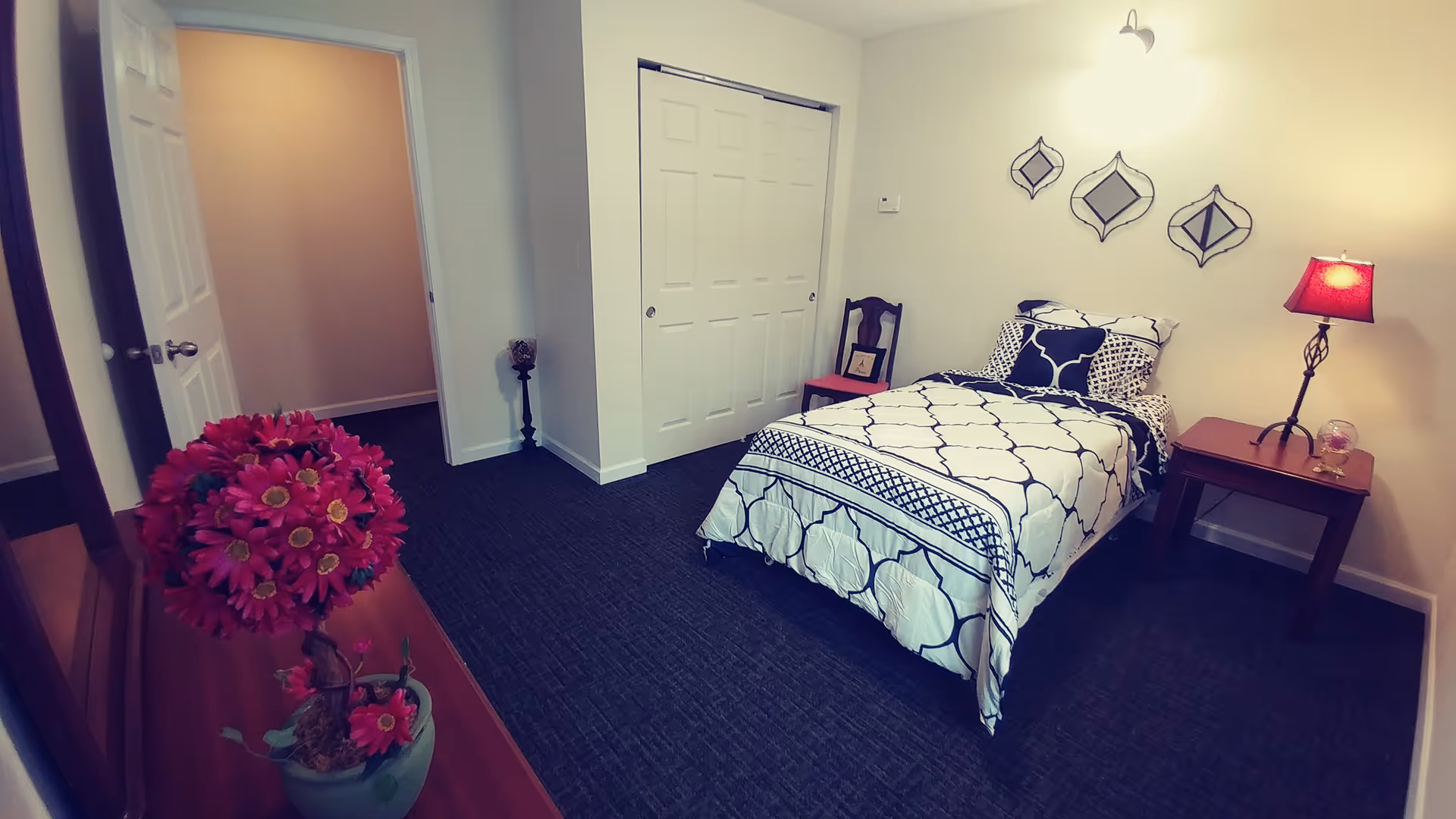 A small bedroom with a single bed covered in a white and black patterned bedspread. There is a wooden nightstand with a red lampshade on the right side of the bed, and a wooden chair with a red cushion next to the bed. Three decorative mirrors are mounted on the wall above the bed. A closet with white sliding doors is visible on the far wall. A wooden dresser with a vase of red flowers is in the foreground on the left side. The room has dark carpet flooring and light-colored walls.