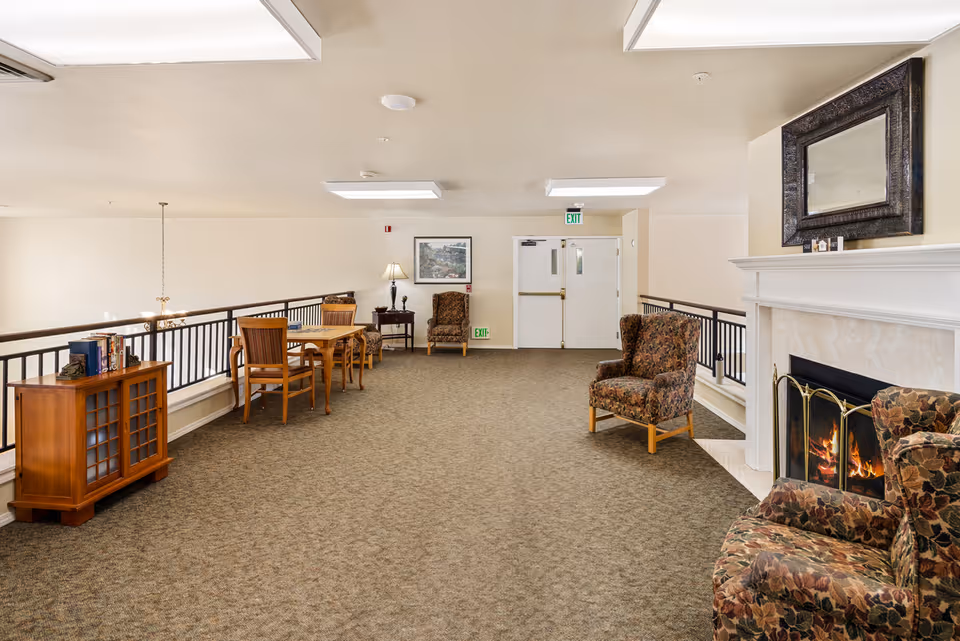 A spacious indoor common area with carpeted floor, featuring a fireplace with a fire burning and two floral upholstered armchairs nearby. There is a wooden table with chairs along the left side, a small cabinet with books, a lamp on a side table, and a framed picture on the wall. Double exit doors are visible at the far end of the room.