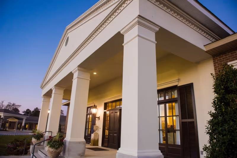 Exterior view of the entrance to Golden Age Healthcare & Rehabilitation Center at dusk, featuring large white columns, double doors with glass panels, outdoor lighting, and surrounding landscaping.