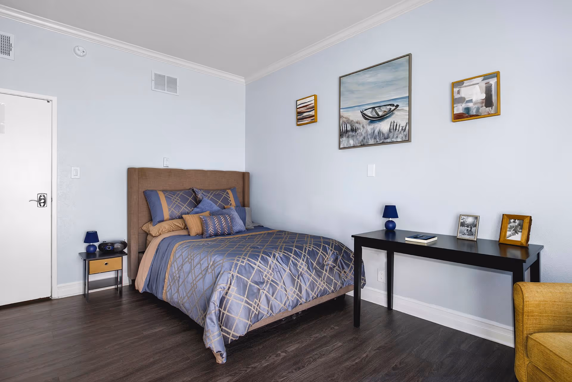 A neatly made bed with blue and beige patterned bedding in a bedroom with light blue walls. There is a small black nightstand with a blue lamp and a radio next to the bed. On the opposite wall, there is a black table with a blue lamp, a book, and three framed photos. Above the table, three framed paintings hang on the wall. The floor is dark wood, and a yellow armchair is partially visible on the right.