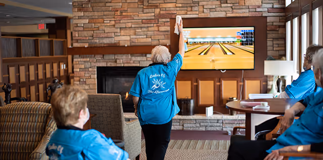 A group of elderly people wearing matching blue shirts are gathered in a cozy living room area with a stone fireplace and a large TV screen displaying a virtual bowling game. One person is standing and appears to be playing the game while others watch seated around tables and chairs.