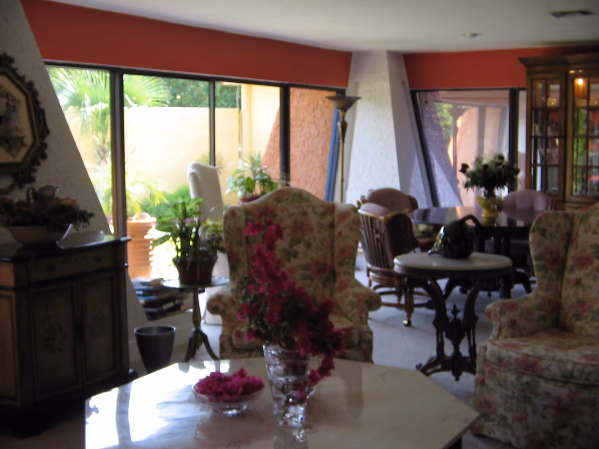 A cozy living room area with floral upholstered armchairs, a marble coffee table with a glass vase of pink flowers, a wooden side table with plants, and a dining table with chairs in the background. Large windows provide a view of greenery outside.