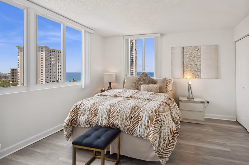 A bright bedroom with large windows showing a view of tall buildings and the ocean. The room features a bed with a patterned comforter and multiple pillows, a small bench at the foot of the bed, a nightstand with a lamp, and a piece of textured wall art above the nightstand.