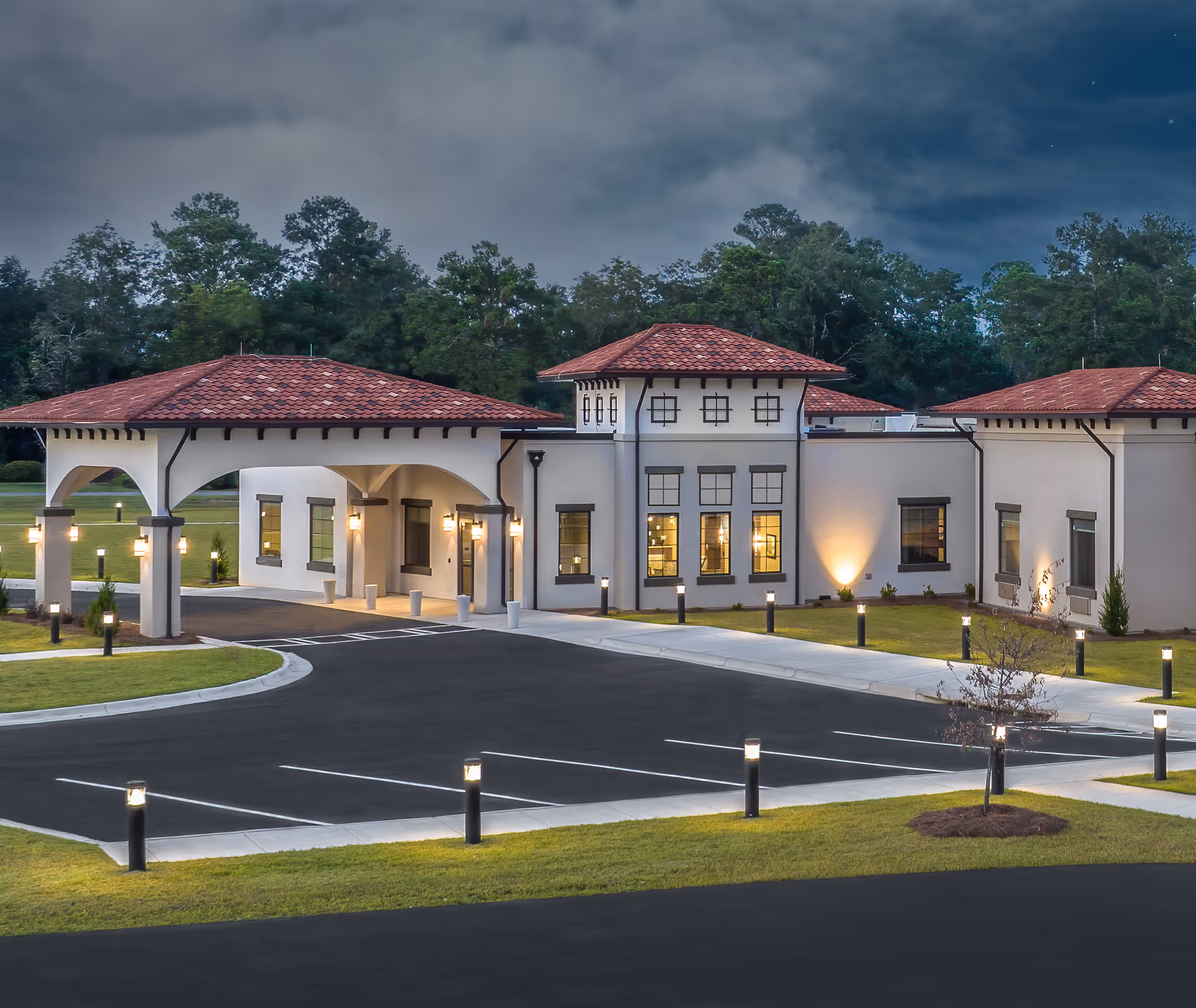 Exterior view of Archbold Living - Cairo facility at dusk, showing a modern building with white walls and red tiled roofs, surrounded by a well-lit parking lot and landscaped greenery with trees in the background.