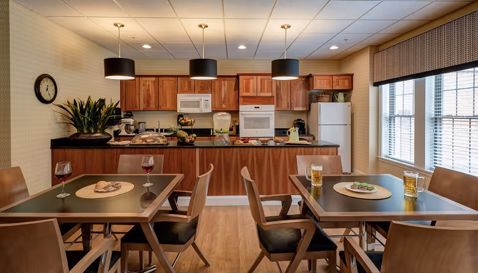 A cozy dining area with two wooden tables, each set with drinks and snacks. The background features a kitchen with wooden cabinets, a white microwave, oven, and refrigerator. Three pendant lights hang from the ceiling, and a large window with blinds allows natural light to fill the room. A wall clock and a plant are also visible.