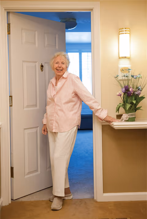 An elderly woman with short white hair, wearing a light pink striped shirt and white pants, stands smiling in a doorway inside a residential facility. The door is open, revealing a room with blue carpet and a window with white blinds. To the right of the door is a small shelf with a vase of purple and white flowers and a wall-mounted light fixture above it.