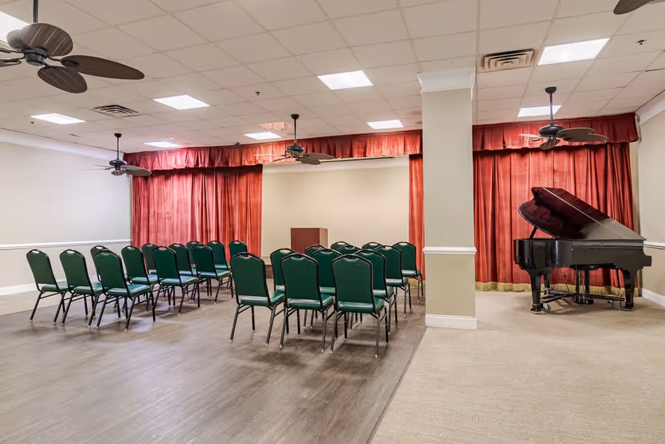 A room set up for a small audience with green chairs arranged in rows facing a podium. The room has a wooden floor section and a carpeted section with a black grand piano on the carpet. Red curtains cover the back wall, and ceiling fans and recessed lights are visible on the ceiling.