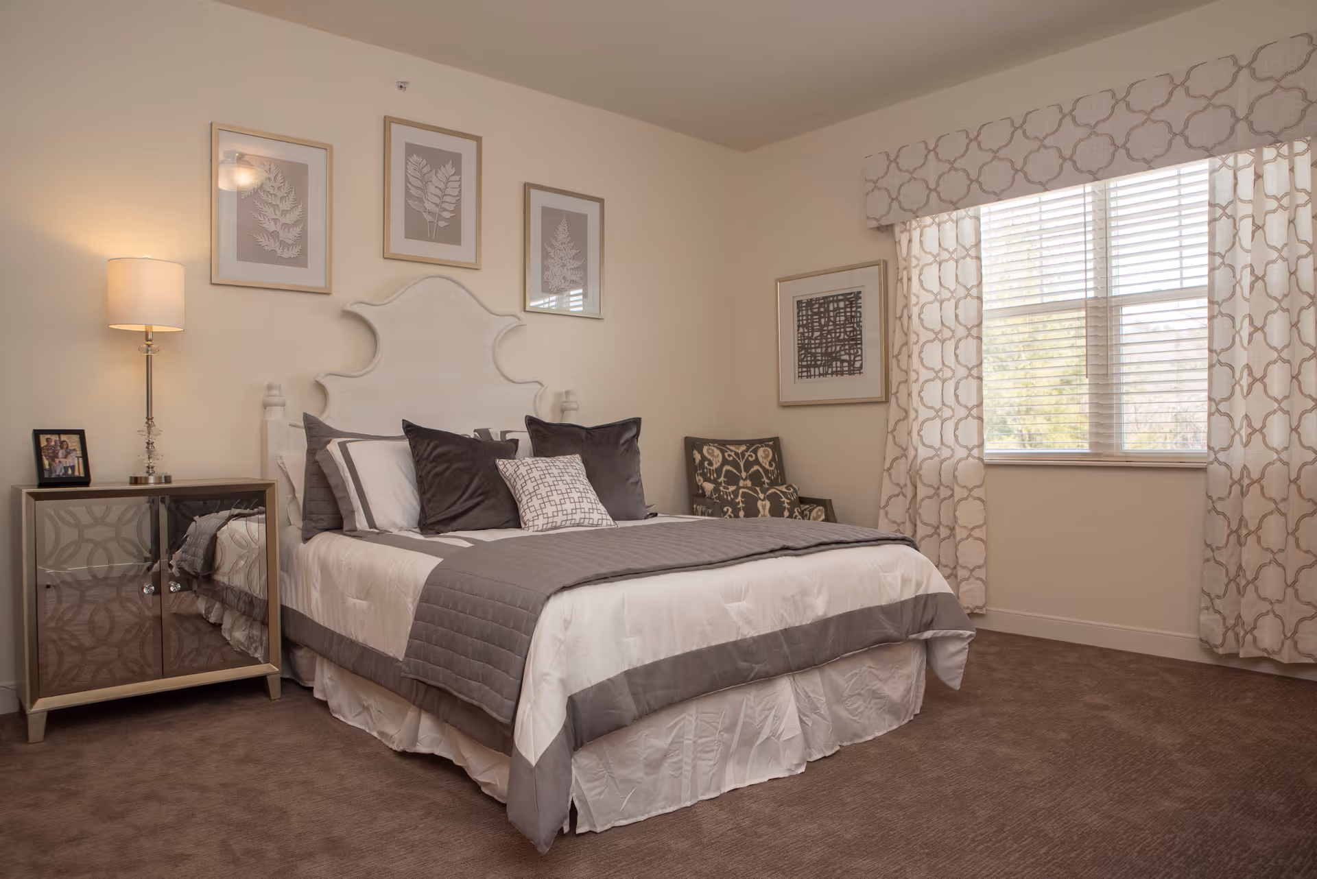 A neatly made bed with white and gray bedding in a bedroom. The bed has a white headboard and several decorative pillows. To the left of the bed is a nightstand with a lamp and a framed photo. On the wall above the bed are three framed botanical prints. To the right of the bed is a patterned armchair and a window with patterned curtains letting in natural light.
