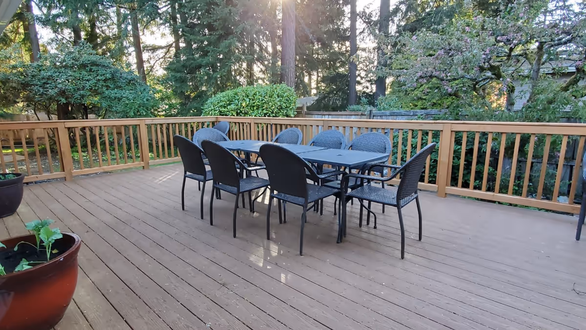Outdoor wooden deck with a black metal table and eight matching chairs surrounded by wooden railing. The deck overlooks a garden area with trees and bushes, with sunlight filtering through the trees.