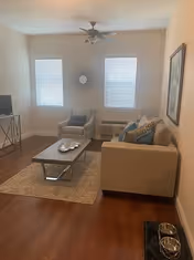 A cozy living room with wooden flooring featuring a beige sofa with blue and beige cushions, a patterned armchair, a wooden coffee table on a beige rug, two windows with blinds, a ceiling fan, a small TV on a stand, a wall mirror between the windows, and a framed picture on the wall.