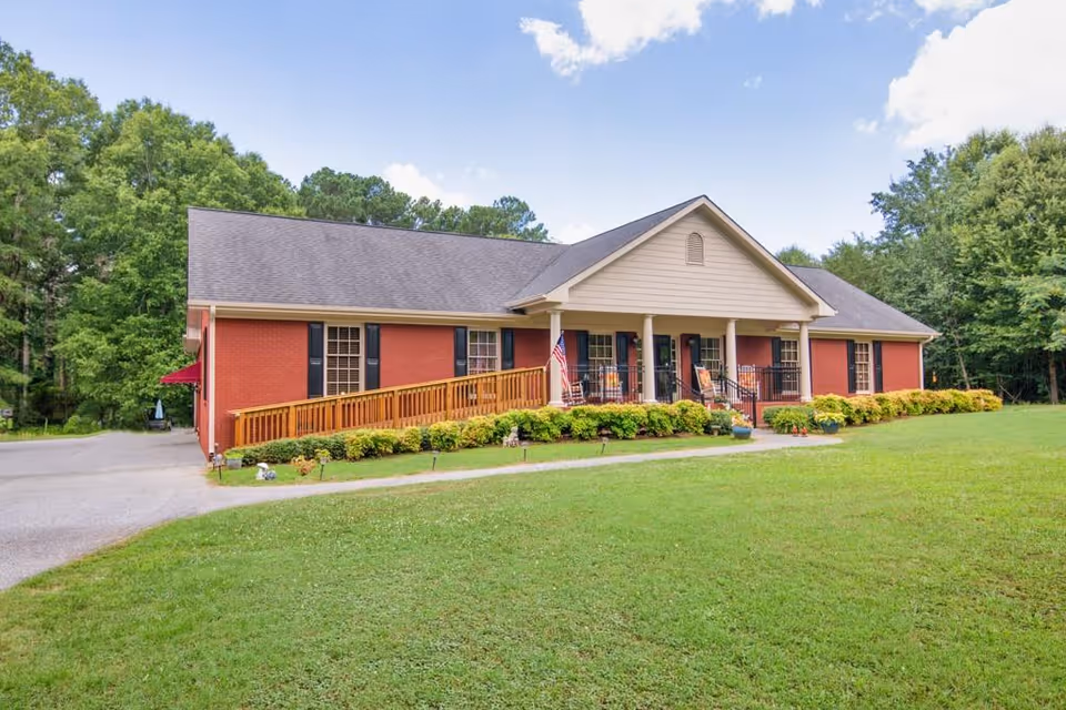 Single-story brick building with a gray shingled roof, a wooden ramp leading to the front porch, and several windows with black shutters. The porch has white columns, rocking chairs, and an American flag. The building is surrounded by green grass, bushes, and trees under a partly cloudy sky.