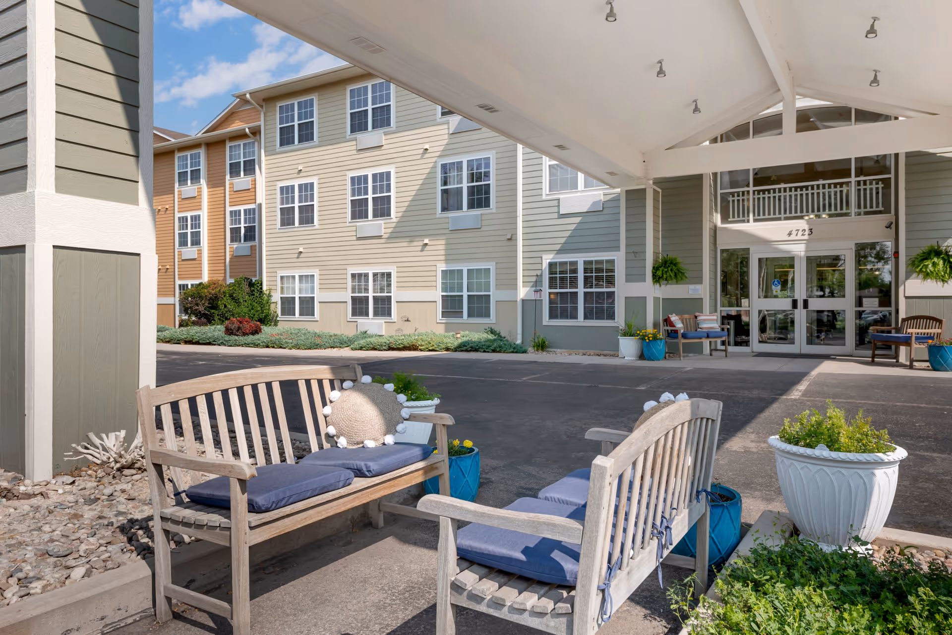 Outdoor seating area with wooden benches featuring blue cushions and decorative pillows under a covered entrance. The entrance leads to a multi-story building with beige and green siding, multiple windows, and the number 4723 above the glass doors. Potted plants and greenery surround the seating area.