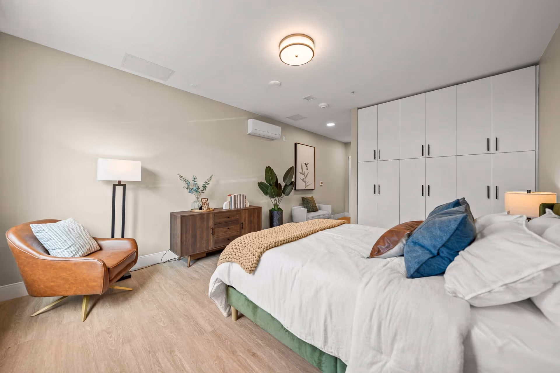 A modern bedroom with a large bed featuring white bedding and multiple pillows. To the right of the bed is a nightstand with a lamp. On the left side of the room, there is a brown leather armchair with a pillow, a floor lamp, and a wooden dresser with books and a framed photo on top. A large plant and a white armchair are positioned near the back wall, which also has a framed botanical print. The room has light wood flooring, beige walls, and a ceiling light fixture. White built-in cabinets with black handles line one wall.