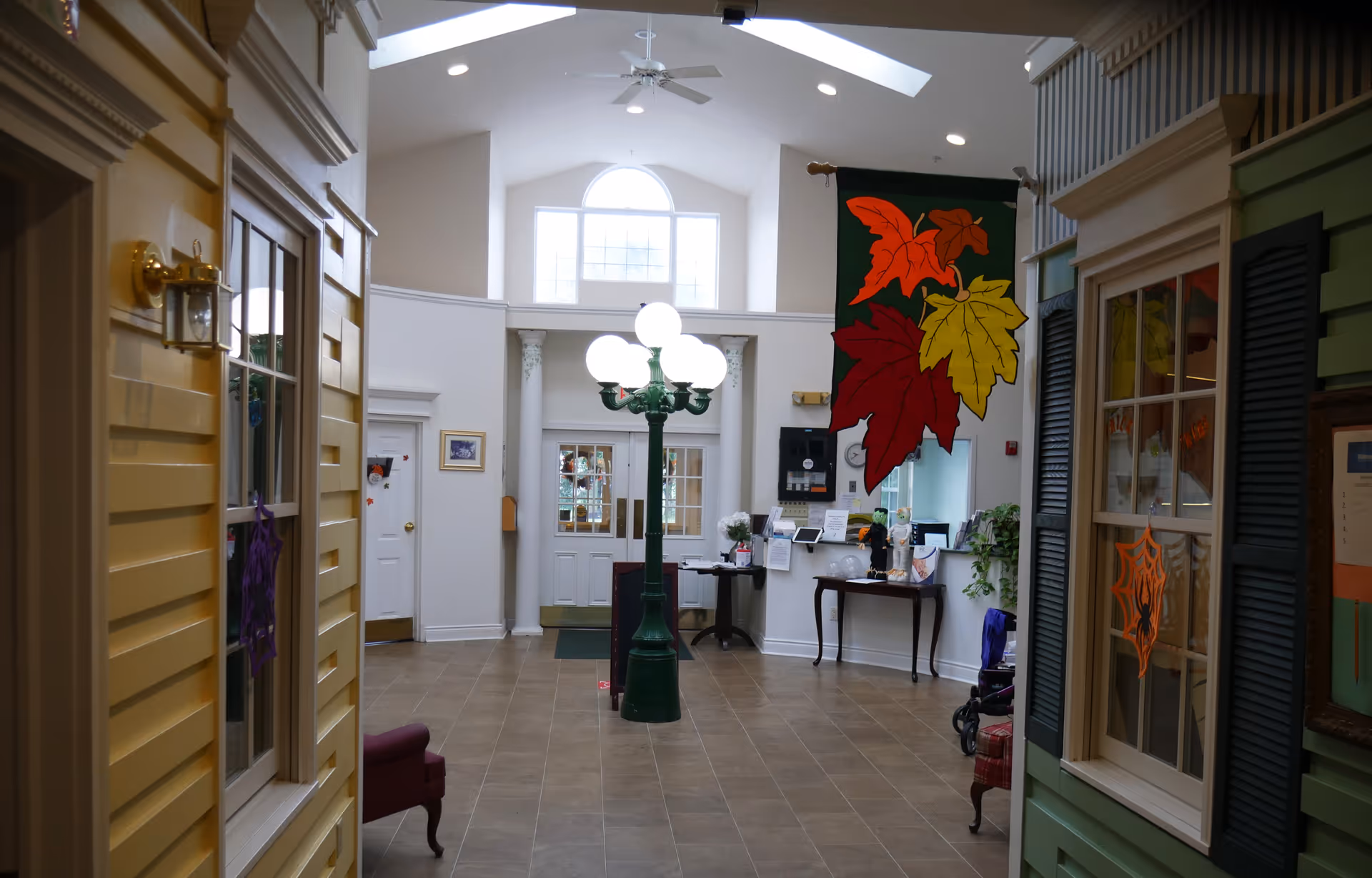 Interior view of a senior living facility hallway with high ceilings and skylights. The hallway features decorative house-like facades on either side, a green lamp post with globe lights in the center, and autumn-themed decorations including large colorful leaves hanging from the ceiling. There are tables with flowers and informational materials near the far wall, and a door with windows letting in natural light.