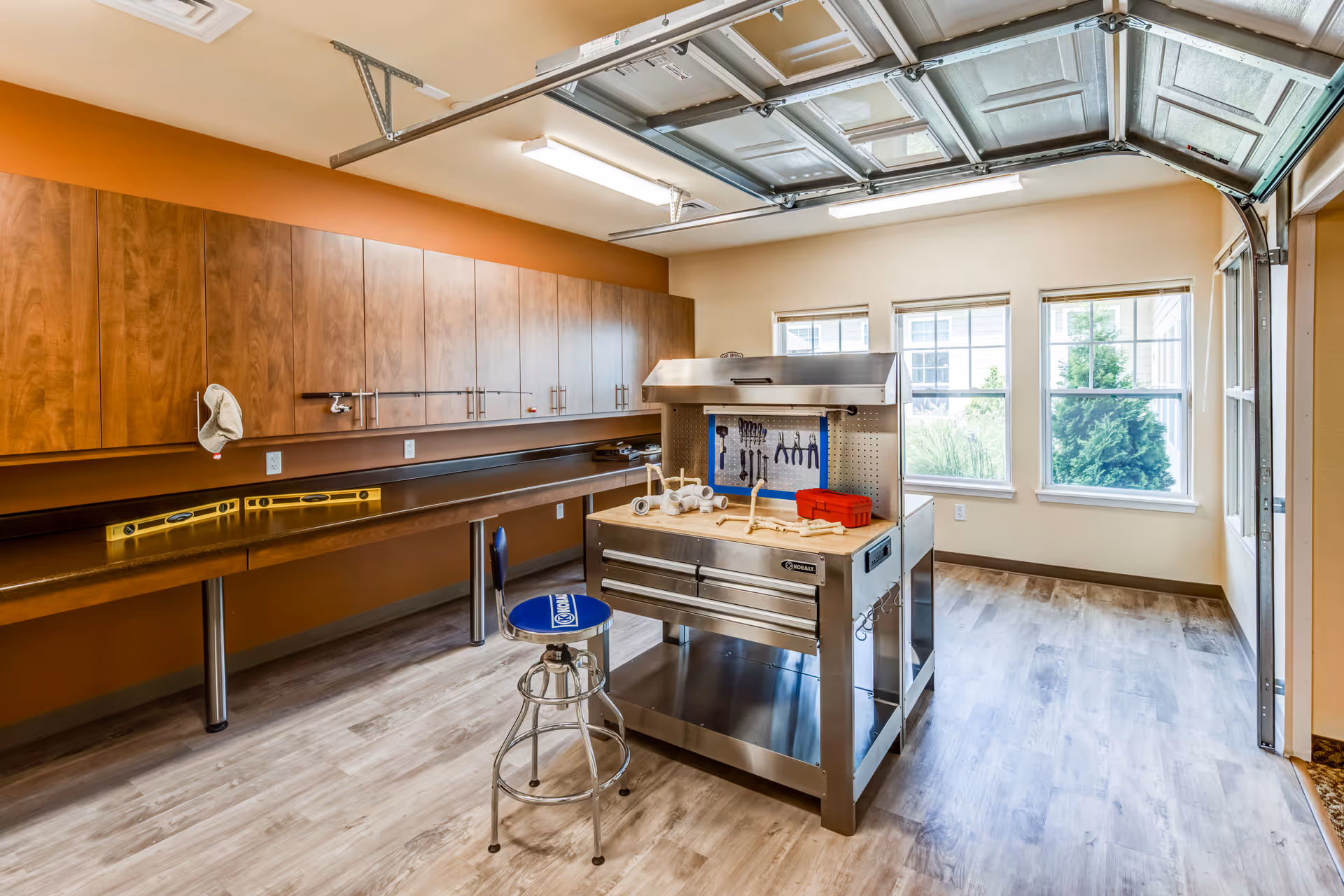 Bright interior workshop with a central stainless steel workbench, tool board, long counter and wall cabinets under a raised garage-style door.