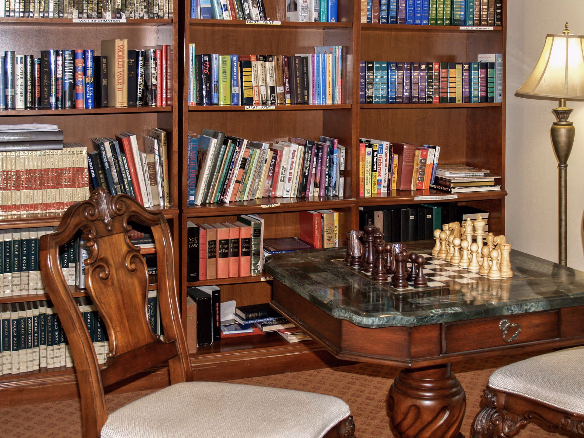 A cozy reading and game area featuring a wooden bookshelf filled with books, a wooden chair with an upholstered seat, a table with a chessboard set up for a game, and a floor lamp providing warm light.