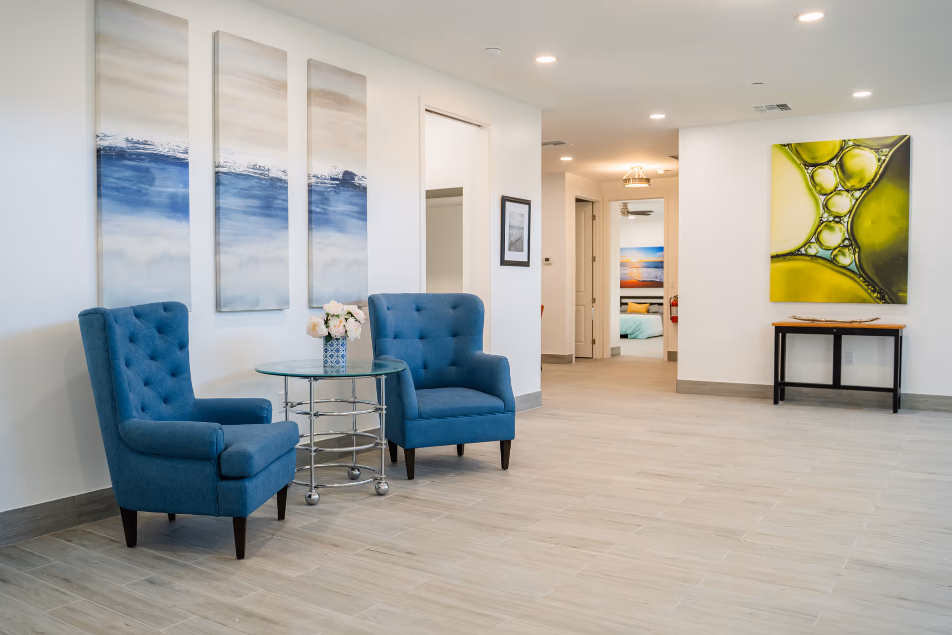 A bright and spacious interior area with two blue upholstered armchairs and a round glass table with a vase of flowers between them. The walls are decorated with abstract paintings, and there is a hallway leading to other rooms visible in the background.