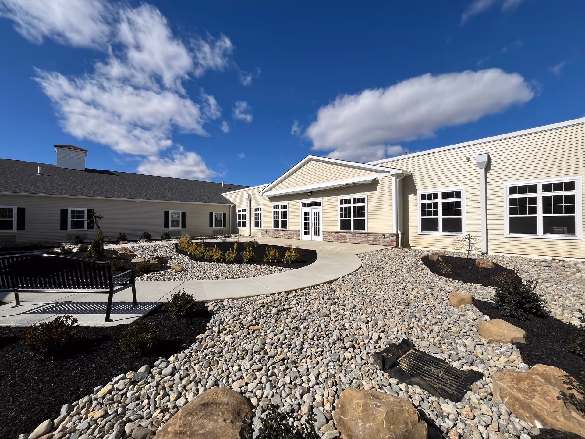 Front courtyard of a single-story building with a paved walkway, bench, rock landscaping, and beige siding under a blue sky.