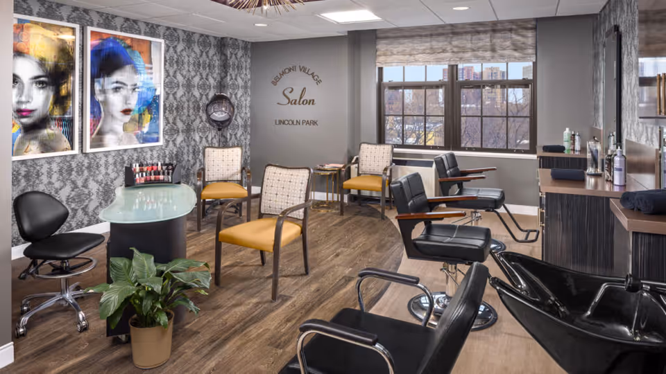 Interior view of Belmont Village Senior Living Lincoln Park salon featuring salon chairs, a manicure table with nail polish, decorative wall art, a plant, and large windows letting in natural light.
