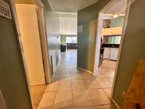 View down a tiled hallway in a senior care home with green walls. To the right, there is an open doorway leading to a kitchen area with light wood cabinets and white appliances. At the end of the hallway, a living room area with a window and a dark-colored couch is visible.