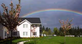 A white two-story building with a dark roof surrounded by green grass and small trees under a cloudy sky with a rainbow arching above.