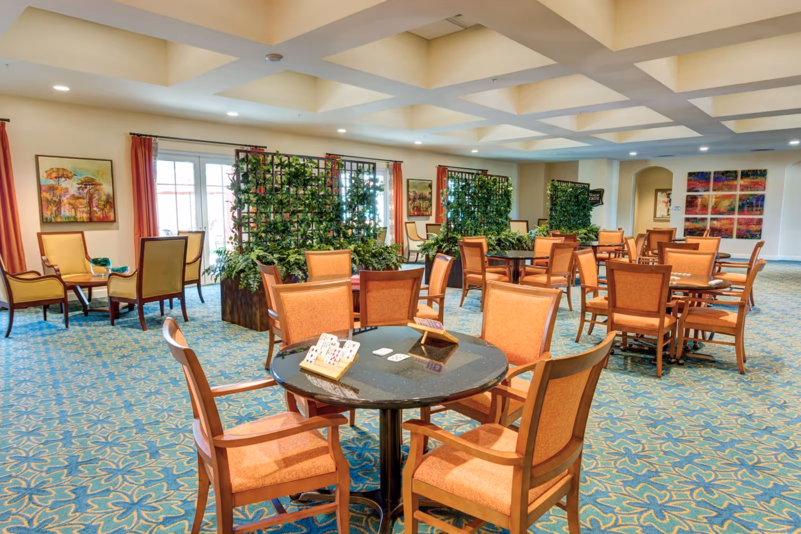 A spacious common area with multiple round tables and orange cushioned chairs arranged for card games, some tables have card holders and playing cards on them. The room features a blue patterned carpet, large windows with red curtains, decorative green plants on trellises, and colorful artwork on the walls. The ceiling has a coffered design with recessed lighting.