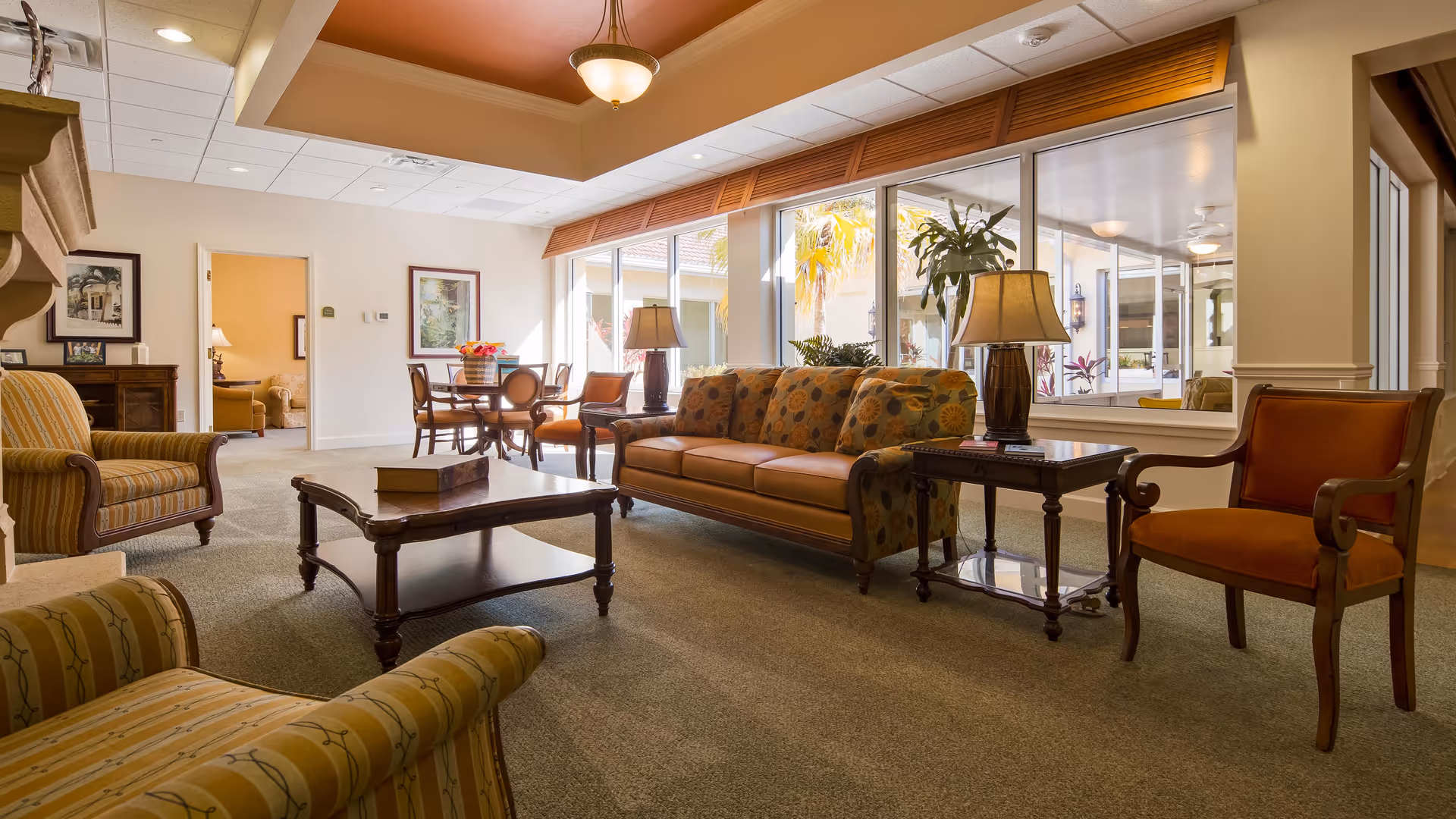 A well-lit living room area in a senior living facility with comfortable upholstered chairs and a sofa arranged around a wooden coffee table. There are two table lamps on side tables, large windows letting in natural light, and a dining table with chairs in the background. The room has a warm and inviting atmosphere with neutral tones and tasteful decor.