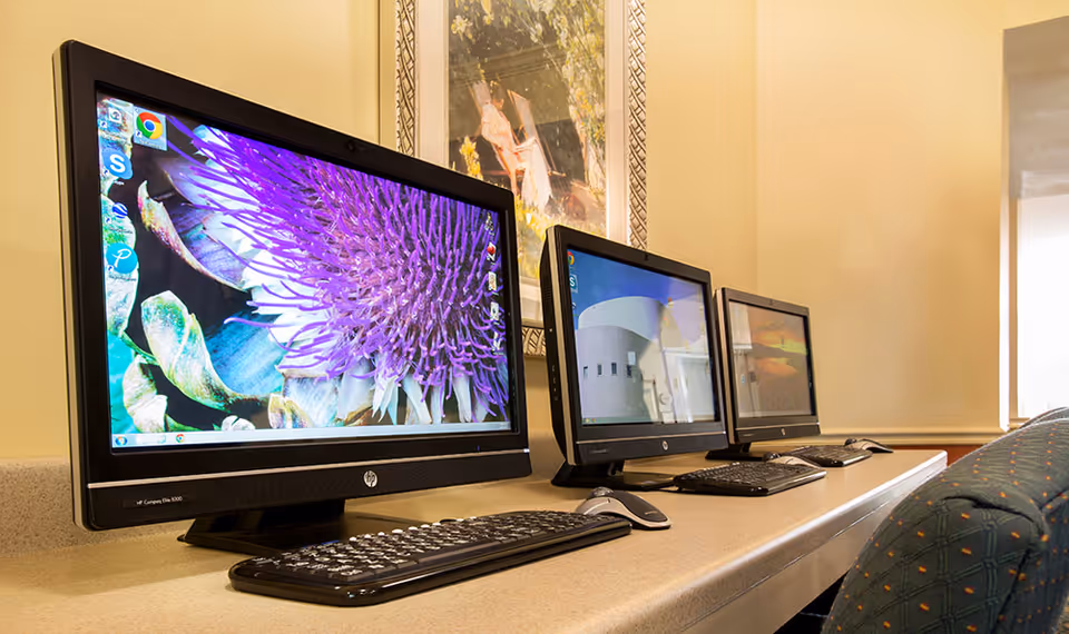 Three desktop computers with keyboards and mice arranged on a long counter in a room with beige walls and a framed picture hanging above.