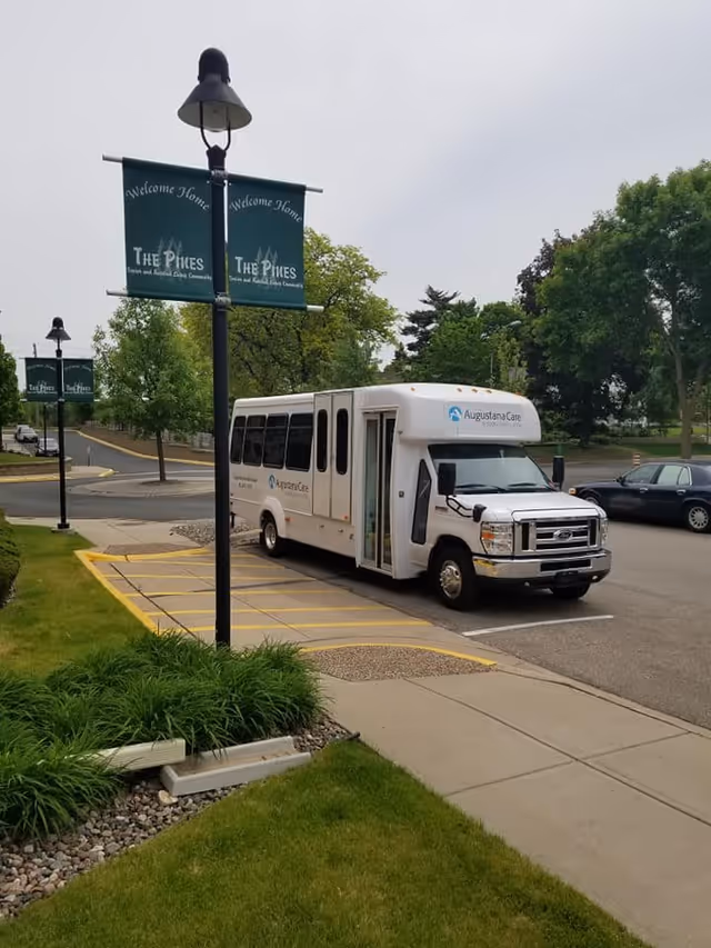 A white shuttle bus parked in a parking lot near a sidewalk with green grass and plants. Several green banners on lamp posts read 'Welcome Home, The Pines Senior and Assisted Living Community'. Trees and other parked cars are visible in the background under an overcast sky.