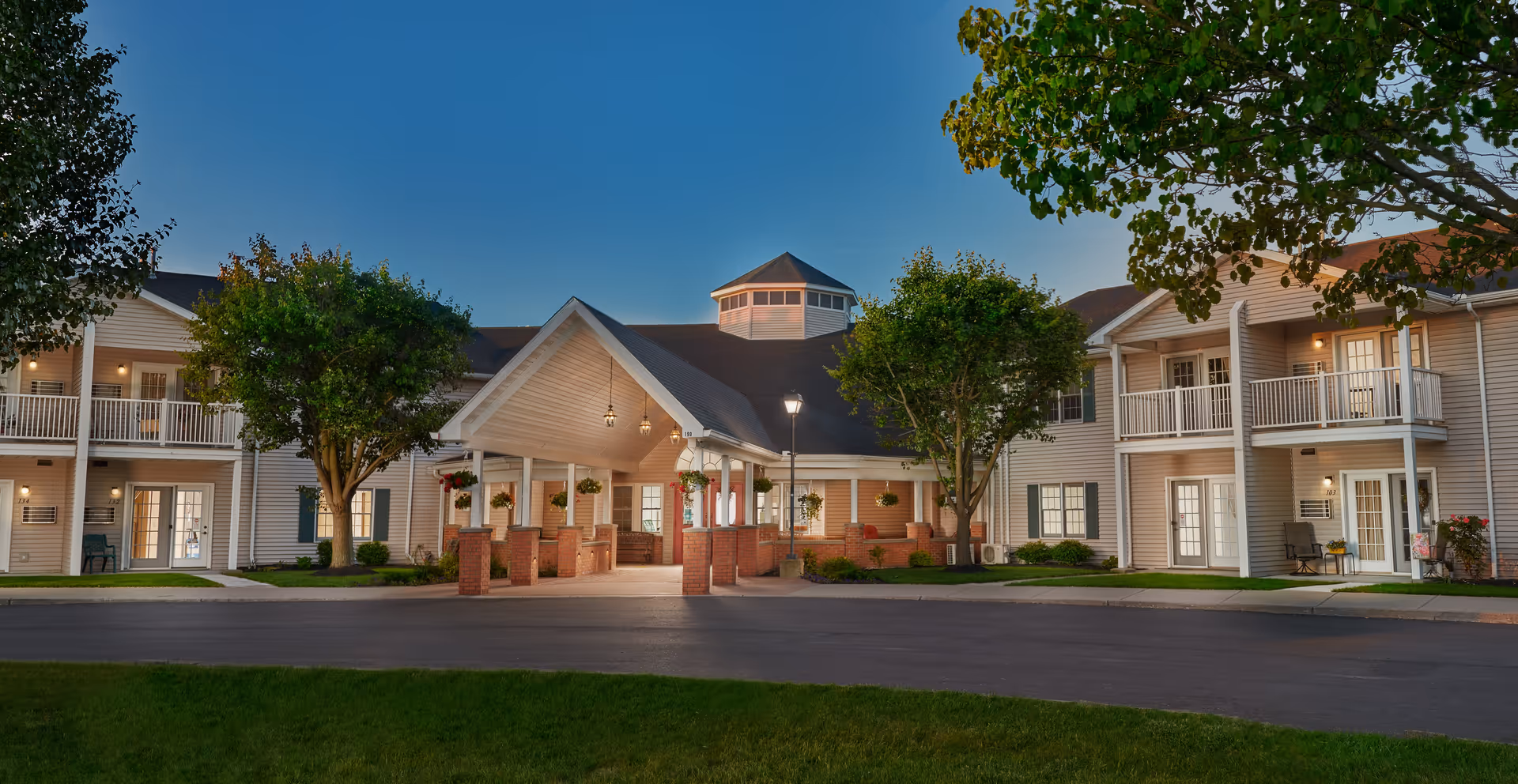 Exterior view of Ashton Place Senior Living facility at dusk, showing a two-story building with balconies, a covered entrance with brick pillars, hanging flower baskets, and well-maintained landscaping including trees and grass.
