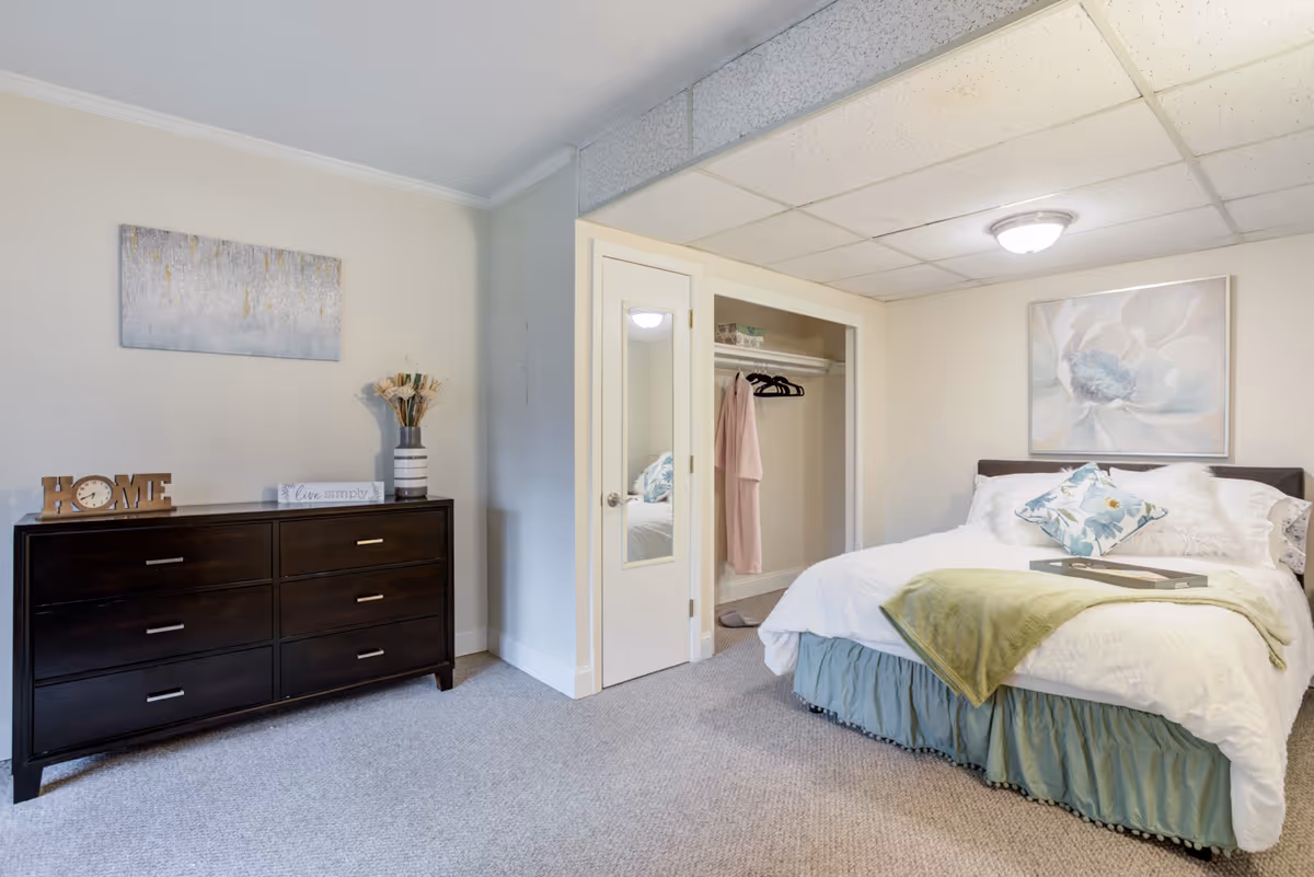 A bright bedroom with a made bed, pillows and throw, a dark dresser with decor, wall art, and an open closet.
