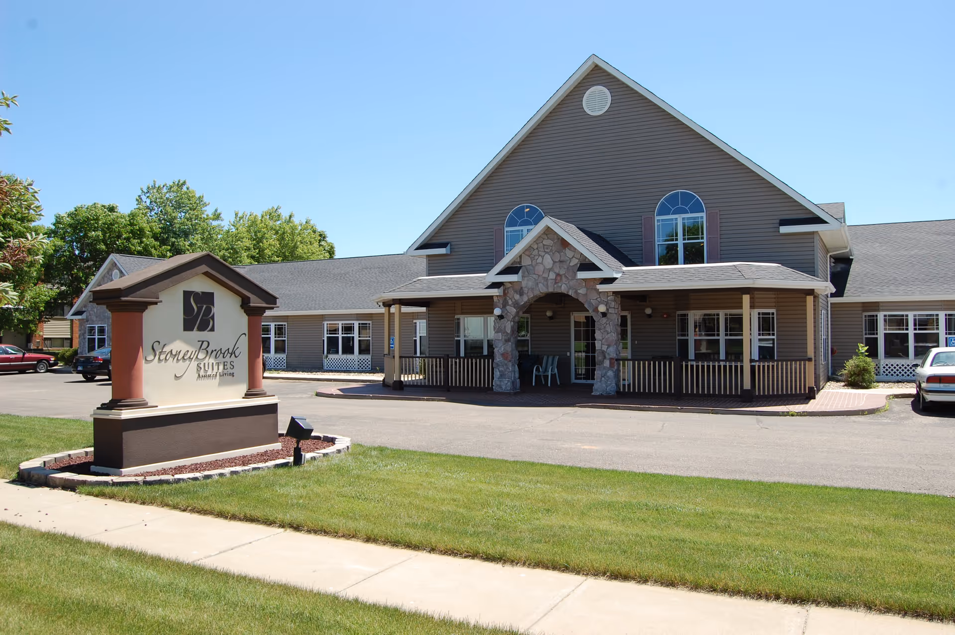 Exterior view of StoneyBrook Suites Assisted Living facility showing a large building with a peaked roof, stone arch entrance, multiple windows, and a parking area with cars. A sign with the facility name is prominently displayed on a landscaped lawn near the sidewalk.