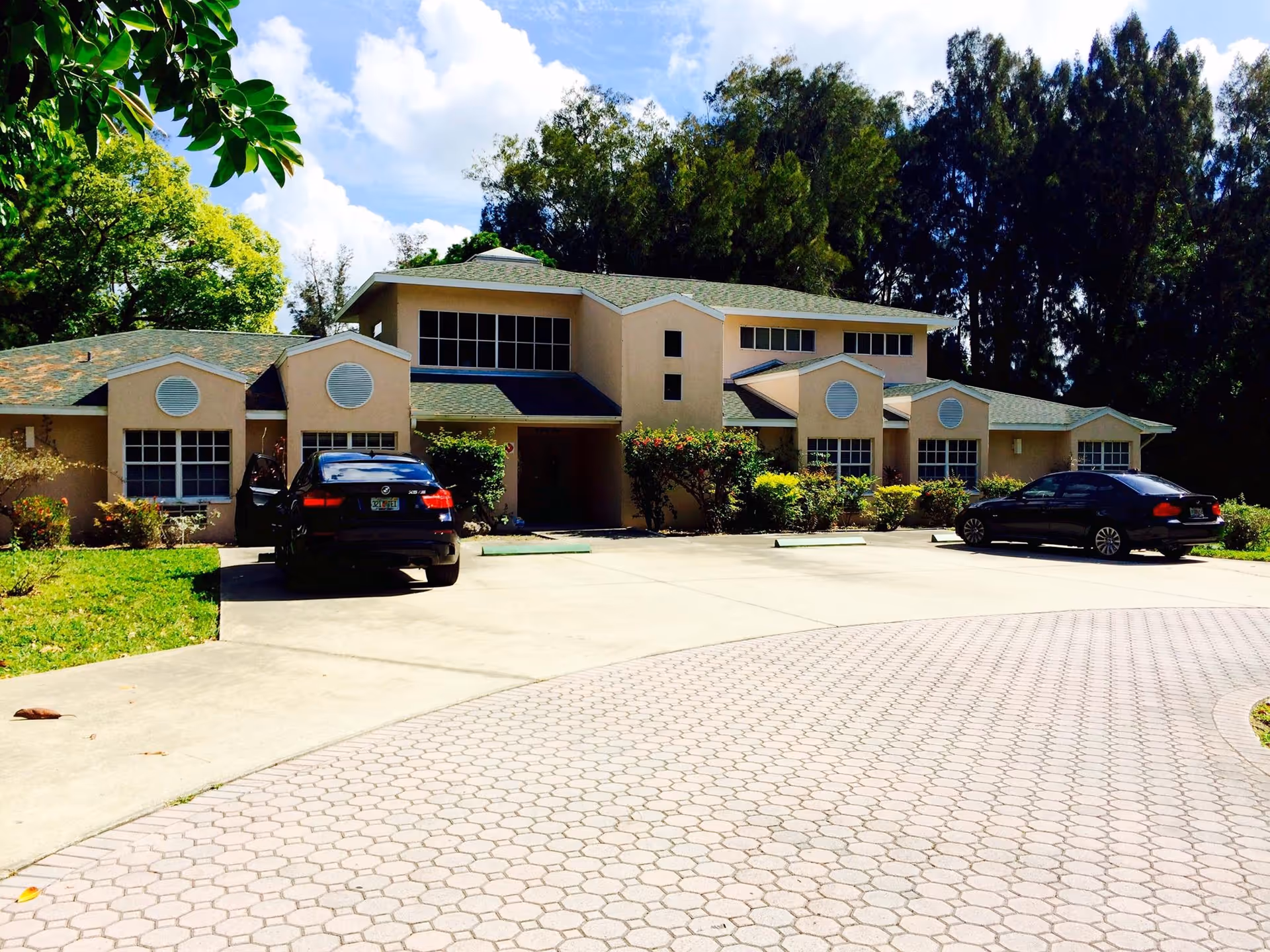 Front exterior of a low-rise beige residential building with two parked cars in the driveway and trees behind it.