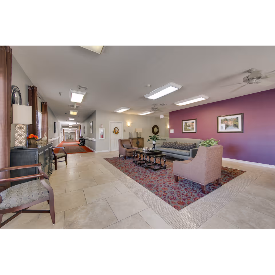 A spacious senior living facility common area with a seating arrangement including a sofa and two armchairs around a coffee table on a patterned rug. The walls are painted purple and beige, decorated with framed artwork. The area is well-lit with ceiling lights and fans. A long hallway extends from the seating area with additional chairs and decor along the walls.