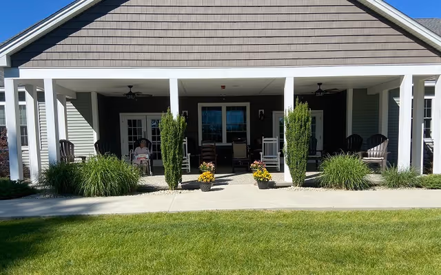 Front porch area of a building with a covered patio featuring white columns, rocking chairs, potted plants, and shrubs. The building has gray siding and a peaked roof. A person is sitting on one of the chairs on the left side of the porch.